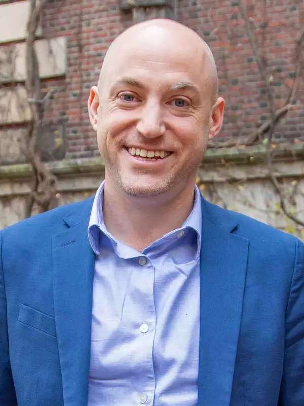 A-J Aronstein smiling in front of a brown building wearing a collared button up shirt (lavender) and a blue blazer