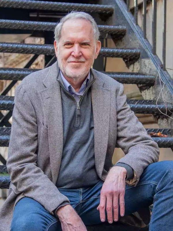 staircase in background, man with gray blazer and blue jeans, close lipped smile