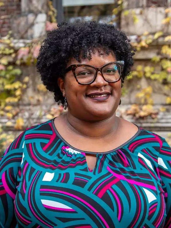 fall background, woman smiling with curly black hair, wearing glasses and a colorful pattered top, smiling